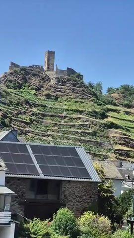 The video shows a view from a moving vehicle, likely a train, looking out at a European town nestled at the base of a steep hill. A castle ruin is visible atop the hill, with terraced vineyards below it. The foreground shows glimpses of houses with traditional European architecture, including some with timber framing and slate roofs. The sky is clear and blue, indicating a sunny day. The camera pans slightly, capturing the passing scenery.