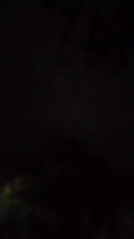 The video displays a dark outdoor scene at night. In the lower left quadrant, a bright outdoor light illuminates a portion of a structure and some foliage. The sky is predominantly black with faint clouds visible. Toward the end of the video, a flash of purple light appears in the sky, suggesting lightning. There are no people or discernible interactions depicted.