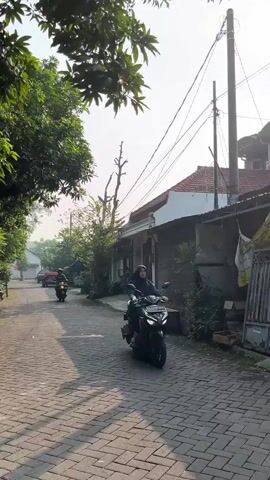 A person rides a dark colored scooter down a narrow paved road lined with trees and houses. The road is made of grey bricks laid in a pattern. On the right side of the road, there is a house with a white facade and a red tiled roof. There are also some plants and a wooden bench next to the house. On the left side of the road, there are more trees and houses, and a red car can be seen parked in the distance. The sky is bright and hazy. The scooter rider is wearing a helmet.