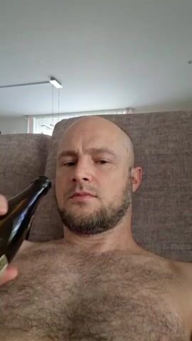 The video begins with a close-up shot of a bald, middle-aged man with a beard drinking from a brown glass bottle. The man is shirtless, revealing a hairy chest. He is lying down, likely on a couch or chair, as indicated by the textured fabric in the background. The lighting is natural, coming from a window or light source off-camera. The man takes a drink from the bottle and then sets it down on his chest. The camera angle is low, as if the person recording is also lying down. The overall environment appears to be a domestic setting.
