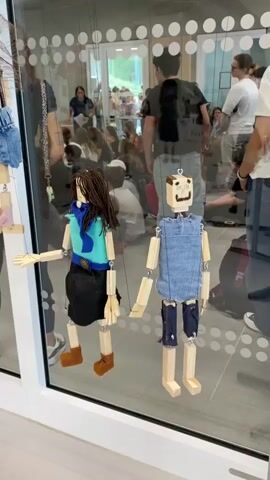 The video shows a display of handmade puppets visible through a large glass window. The puppets are made of wood and fabric, with some featuring yarn for hair and denim for clothing. They are hanging at various heights, some appearing to be suspended by strings. Behind the puppets, through the glass, a group of people are gathered in what appears to be an indoor space. Some individuals are seated on the floor, while others are standing. The environment suggests a classroom or exhibition setting, possibly a school or art event, given the presence of multiple people and the displayed artwork. The lighting is bright and even, illuminating both the puppets and the background activity. The perspective is from outside the glass, looking into the room.
