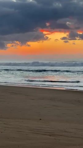 A video shows a wide shot of a beach with a sunset in the background. The sand in the foreground is a light brown color with some ripples. The ocean is in the middle ground with waves rolling onto the shore. The sky in the background is filled with dark clouds and a vibrant orange and red sunset. There are no people or objects visible in the video.
