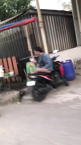 A person is sitting on a red and black motorcycle. They are wearing a blue shirt and tan shorts. The motorcycle is parked on a paved area next to a building with corrugated metal siding. There is a wooden fence to the left of the motorcycle, and a blue barrel to the right. The person appears to be holding a white object in their hands. The video is blurry and appears to be shot from a moving vehicle.