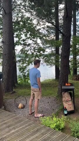 A man wearing a blue t-shirt and tan shorts stands in front of a hot tub on a wooden deck. He is facing away from the camera, looking out at a lake. To his right, a barbecue grill with a fire burning is visible. There is a pile of logs next to the grill. The hot tub is made of wood and has a black cover. The deck extends to the edge of the lake, which is surrounded by trees and vegetation. The sky is cloudy with a hint of sunset colors. In the background, a hot tub chimney is visible. To the left of the frame, a portion of a hot tub is seen. The video pans from the man and the hot tub to a wider view of the lake and surrounding landscape. A pool net and pole are visible near the edge of the deck.