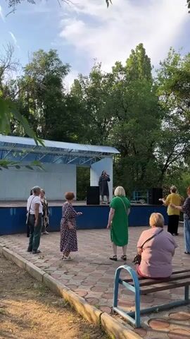 A group of elderly people are gathered outdoors near a stage. A woman in a dark dress stands on the stage, holding a microphone and appearing to sing or speak. Several other individuals, mostly women, are positioned on the paved area in front of the stage, some standing and some seated on a blue bench. They are facing the stage. Trees and foliage form the background, suggesting a park or outdoor venue. The lighting indicates it is daytime, possibly late afternoon.