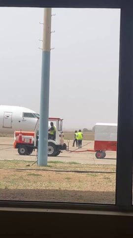 The scene captures a Value Jet aircraft on the tarmac, viewed through a window, possibly at an airport. The aircraft is white with the company logo clearly visible. In the foreground, there are two SAHCO PLC baggage carts, one connected to a small white tractor. A worker wearing a yellow vest is driving the tractor, and another worker in similar attire is standing near the carts. The sky appears hazy. The foreground includes some grass and a section of the window frame. A utility pole is also visible.