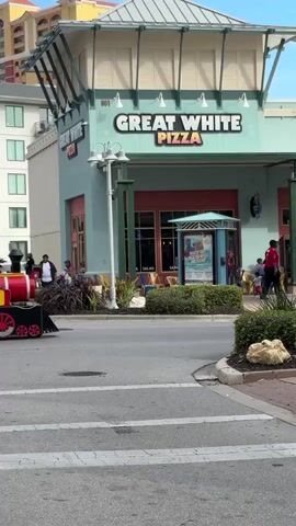 A miniature train with a red engine and several passenger cars travels down a street in front of a building with a sign that reads "GREAT WHITE PIZZA." The train is carrying passengers. People are visible on the street and sidewalks, and there are buildings in the background. The weather appears to be sunny.