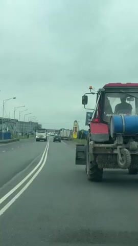 The video shows a view from a vehicle driving on a paved road. In the foreground on the right is the back of a red tractor with a blue barrel attached to it and a large brush underneath. In the distance, on the road ahead, are several other vehicles including a white truck and a dark-colored car. On the left side of the road are streetlights and buildings. The sky is overcast. The camera is moving forward and slightly to the left, following the tractor.