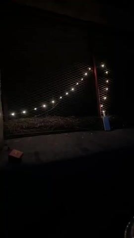 The video shows a dark outdoor scene at night. A chain-link fence runs horizontally across the frame, with a vertical support on the right. A string of small, glowing lights is draped along the fence, forming a diagonal line from left to right and then vertically down the support. Below the fence, there is a pile of dry vegetation or straw. On the ground in front of the fence, a small red rectangular object is visible on the left, and a blue rectangular object, possibly a lighter, is placed near the base of the vertical fence support on the right. The ground is a dark, rough surface. The overall lighting is dim, with the string lights providing the primary illumination.