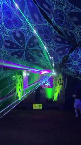 The video shows an outdoor event at night, likely a music festival or party, characterized by vibrant laser light shows and decorations. A canopy, adorned with intricate geometric patterns in blue and green, stretches overhead, serving as a ceiling. Beneath the canopy, a stage area is set up with two figures, possibly DJs, behind a DJ booth. The booth is lit with green and pink lights, and a large, illuminated tree graphic is displayed as a backdrop. Green and purple laser beams cut through the darkness, projecting onto the canopy, the stage, and into the space in front of the booth. To the left and right of the stage are large, cutout figures with illuminated green outlines, adding to the psychedelic atmosphere. A small yellow sign with text in Russian, "ЭТА МУЗЫКА САМА СЕБЯ НЕ ЗАТАНЦУЕТ" (This music will not dance by itself), is visible in front of the DJ booth. The ground appears to be dark and unpaved, with some scattered pink lights creating decorative patterns on the ground in the foreground. A person's legs and feet, wearing a light-colored garment, are visible on the far right edge of the frame, suggesting an audience member. The overall impression is one of an energetic and visually stimulating nocturnal gathering.