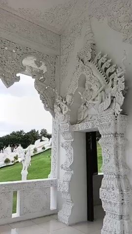 The video pans across a white, ornate structure with intricate carvings. The camera moves from right to left, revealing a balcony with a white railing. Beyond the railing, a green, grassy hillside is visible, dotted with white statues. In the distance, trees and mountains can be seen under a cloudy sky. The overall impression is of a serene and visually striking location, likely a temple or a similar religious or artistic site.
