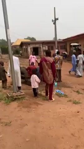 The video shows a group of people gathered in a dusty, open area. Several buildings are visible in the background. A pole with two speakers is located near the center of the frame. The people are dressed in a variety of clothing, including traditional robes and casual wear. Some are standing, others are sitting, and a few are walking around. A person in a red jersey carries a white bucket on their head. A water tank is visible near one of the buildings. Several pairs of shoes are piled up near a doorway. The overall atmosphere appears relaxed and communal.