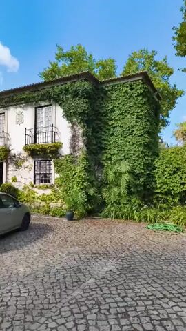 A silver car is parked on a cobblestone driveway in front of a white building covered in ivy. The building has several windows and balconies. Tall green trees surround the building and driveway. The sky is blue with scattered white clouds.