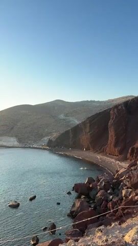 The video captures a scenic view of a bay at sunset. The camera pans across the water, revealing several sailboats and a few smaller boats anchored or moving slowly. The sun is low on the horizon, casting a warm, golden glow on the water. The surrounding landscape consists of rolling, barren hills with some roads winding up their slopes. On the right side of the frame, a steep, reddish-brown cliff face is visible. The water is calm with gentle ripples, and some rocks are scattered along the shoreline in the foreground. The sky is clear and a soft blue, gradually transitioning to a lighter hue near the horizon. The overall atmosphere is peaceful and tranquil, characteristic of a serene coastal sunset.