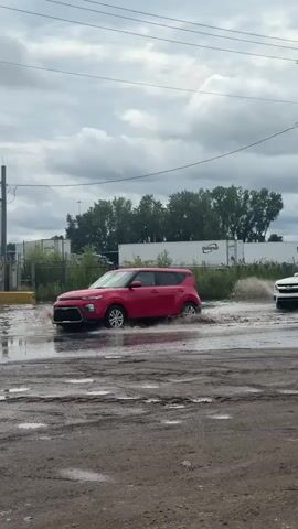A red Kia Soul drives through a flooded area, creating a splash. A white pickup truck with a logo that says "vivint SmartHome" follows, also driving through the water. A white van with logos that say "Toyota" drives past. The sky is cloudy. The ground is muddy and appears to be under construction. There are power lines and poles visible in the background, along with trees and some buildings.