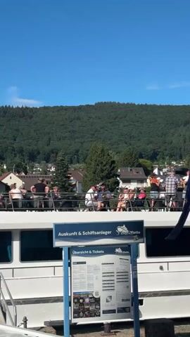 A bright, sunny day with a clear blue sky and a dense, green hillside in the background. In the foreground, the upper deck of a white boat is visible, populated with people. They are seated at tables and standing, some appearing to be tourists. A blue and white sign with German text is mounted on the side of the boat, reading "Auskunft & Schiffsanleger" and displaying information. The people on deck are casually dressed, with a mix of men and women of various ages. Some are talking, some are looking out at the scenery, and one woman is holding a child. The overall atmosphere appears relaxed and leisurely.