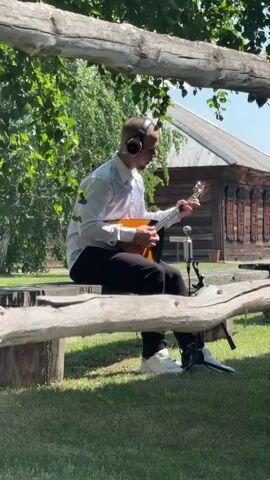 A man wearing headphones and a white dress shirt is sitting on a wooden bench outside and playing a balalaika. He is wearing black pants and white sneakers. A microphone is positioned in front of him. Behind the man, there is a rustic wooden building with decorative trim. The scene is set in a grassy area with trees and foliage. The video is shot from a low angle, looking through natural wooden framing.