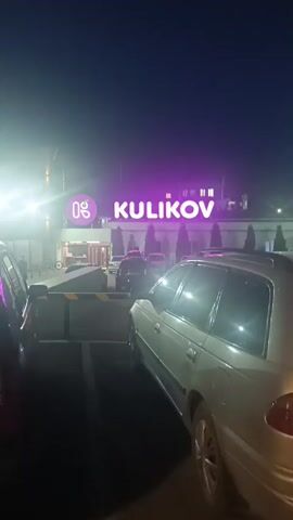 The video shows a nighttime scene in a parking lot in front of a building. The building has a large, bright purple sign that reads "KULIKOV" with a circular logo to its left. There are cars parked in the lot, and some people can be seen walking in front of the building. The lighting suggests it is after dark, and the sky is clear and dark blue. The overall atmosphere appears calm and ordinary, with no specific event clearly indicated. The location is identified as Alamüdün, Kyrgyzstan.
