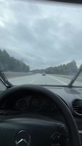 The video shows a first-person perspective of driving on a highway on a cloudy day. The driver is behind the steering wheel of a Mercedes-Benz, and the dashboard is visible. The windshield wipers are on, clearing rain from the glass. Other cars are on the road, and trees line the sides of the highway.