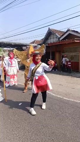 The video shows a parade on a street. In the foreground, a woman in a white and red costume with large wings on her back and a red headscarf is dancing. She has a sash around her neck that reads "DSN CANGKRINGAN". To her left, another person is dressed in an elaborate white ruffled costume, holding a long stick with a large Indonesian flag attached to it. Behind them, other people in similar costumes are walking in the parade. The background shows houses, power lines, and a clear sky. The overall scene appears to be a festive cultural event.