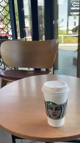 The video shows a close-up of a Starbucks cup on a round wooden table. The cup has a white lid and a cardboard sleeve with the Starbucks logo. In the background, there are wooden chairs and tables, as well as a window looking out onto a street. The overall scene appears to be inside a Starbucks cafe.