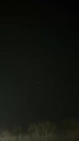 The video shows a dark night sky with the faint glow of lights in the distance. A line of trees is visible along the horizon. A child's voice can be heard in the background. The focus is on the dark sky and distant lights.