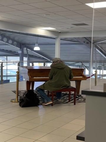 A person is seated at a wooden upright piano, viewed from behind, in what appears to be a bright, modern indoor public space, possibly an airport or transportation hub in Rochford, United Kingdom. The individual wears a long, hooded green coat, blue jeans, and light-colored shoes. A black duffel bag rests on the tiled floor beside a dark-colored piano bench. The piano itself sits on a black and white patterned rug. To the left of the piano, two gold stanchions with red ropes delineate an area. The background features high ceilings with exposed metal framework, large windows or glass partitions, and numerous overhead lights. A transparent protective screen or divider is visible on the right side of the frame. The person is actively playing the piano. The lighting suggests it is daytime. No discernible text is visible in the image.