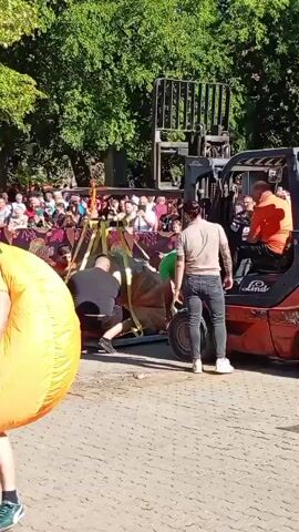 In a sunlit outdoor scene, a man in a large, inflatable pumpkin costume, complete with a carved jack-o'-lantern face, stands in the foreground, facing left. Behind him, a crowd of spectators lines a paved area. In the middle ground, a forklift, operated by a person in an orange shirt, is lifting a large, brown, sack-like object suspended by yellow straps. Several individuals are gathered around the object and the forklift, appearing to be involved in moving or positioning it. The environment includes trees with green foliage and what appears to be a building or structure in the background. The overall atmosphere suggests a public event or festival.