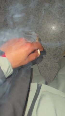 A person's hand is holding a lit cigarette with smoke billowing out of it. The camera angle lowers to reveal the person's leg, clad in light green pants and a multi-toned sneaker. The person then rubs the top of the sneaker with their other hand, which has a red smartwatch on the wrist. The background appears to be a textured, possibly granite, surface.