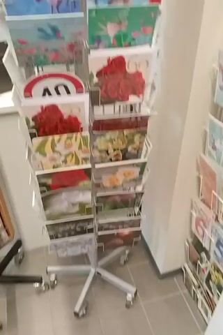 The video shows a close-up, slightly blurry, view of a display rack filled with postcards. The rack is white wire and appears to be on wheels. The postcards are arranged in rows and columns, with a variety of images on them. Some of the postcards feature nature scenes, such as landscapes, animals (deer, foxes, birds, bears), and flowers. There are also some postcards with more abstract or decorative designs, including one with a red rose motif and another with a traffic sign indicating "40". The lighting is typical of an indoor retail environment. The background is mostly out of focus, but appears to be a store interior with other displays and walls visible. The camera is mostly stationary, with some minor panning and tilting. The video quality is poor, with low resolution and motion blur, making it difficult to discern fine details. No people are visible in the video. The video seems to be focused on showcasing the variety of postcards available.