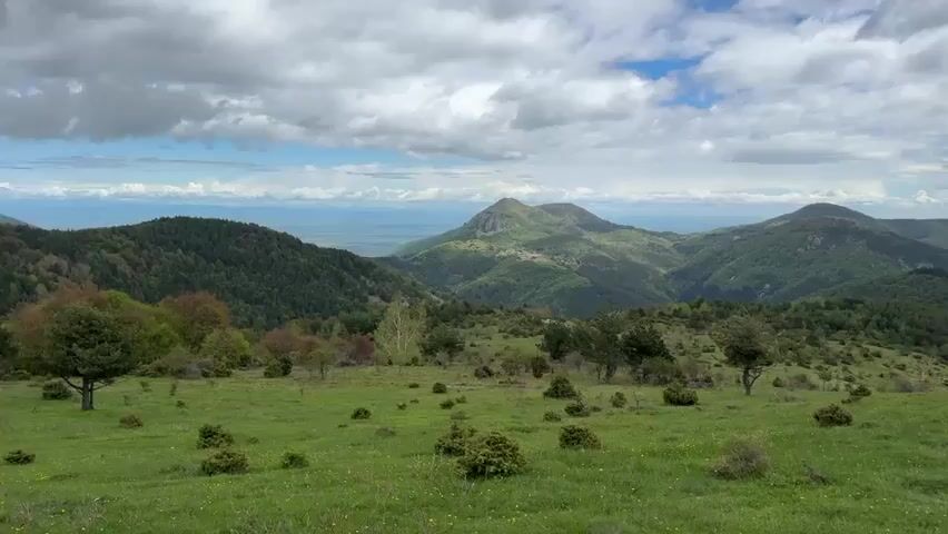The video shows a wide-angle shot of a mountainous landscape. Rolling green hills covered with sparse trees and shrubbery fill the foreground. In the distance, several larger, forested mountains rise towards a cloudy sky. Patches of blue sky are visible between the clouds.  The overall impression is one of a natural, somewhat remote, and peaceful setting.