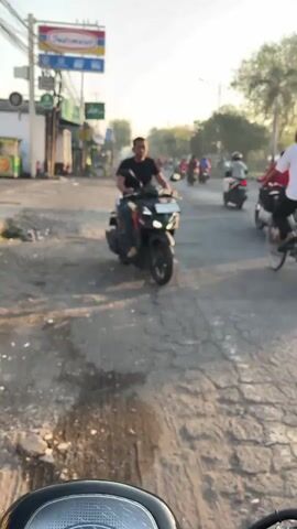 A man on a black scooter is riding on a paved road. The road is in poor condition with many cracks and potholes. There are other people on scooters and bicycles also riding on the road. There are buildings on the left side of the road, including a store with an Indomaret sign. There are trees and streetlights visible in the background. The sky is hazy and appears to be either sunrise or sunset.