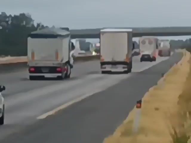 The video shows a view from a vehicle traveling on a highway. The road is paved and has multiple lanes. Several trucks are visible on the highway, some in front and some beside the camera's vehicle. A small white car is also in front of the camera's vehicle. The sky appears overcast. There are trees and vegetation along the side of the road, and an overpass is visible in the distance. The overall impression is of a traffic scene on a cloudy day.