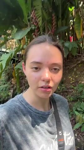 This video compiles several short clips, primarily a selfie video, featuring a young woman in a lush, tropical outdoor environment. The setting is characterized by dense green foliage, large trees, and visible banana plants with bunches of fruit. The time of day appears to be sunny daylight.

In the initial clip, the woman, wearing a grey t-shirt with partially visible text "Easta...", and a black cross-body strap, looks at the camera while moving through the greenery. Her expression is engaged and pleasant.

Around the 0:01 mark, a second woman joins her for a quick selfie. This woman is seen crouching slightly, wearing a green t-shirt with the text "ARQUITECTXS" and a baseball cap, holding a small white notebook. Both women are smiling brightly at the camera.

The video then returns to the primary subject as she continues to film herself walking, her expression consistently cheerful and happy. The background shifts slightly to more clearly showcase tall banana trees and various tropical plants. The overall atmosphere is lighthearted and positive, with both subjects appearing to enjoy their time outdoors.

Based on the provided context, the video was filmed in Pinahuista, Mexico, which aligns with the tropical vegetation seen throughout the clips.