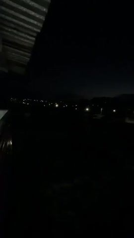 The video captures a dark outdoor scene at night. In the foreground, the edge of a corrugated metal roof is visible on the left. The middle ground is largely obscured by darkness, but a patch of light snow or white material is discernible. The background features a dimly lit rural landscape with scattered streetlights and the silhouette of distant hills or mountains against a dark sky. The lighting is minimal, originating from artificial sources like streetlights and possibly lights within unseen buildings. No people or animals are visible in the frame. The overall impression is one of a quiet, undeveloped area at night.