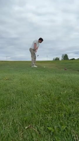 A man wearing khaki pants and a light-colored polo shirt is on a golf course. He has just swung a golf club and is watching where the ball goes. The golf course has green grass and a partly cloudy sky. There is a golf flag in the distance and another person visible farther away.