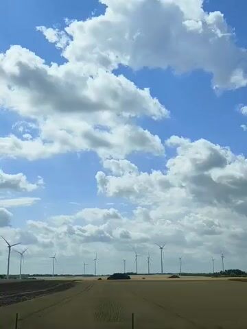 This video shows a landscape with a bright blue sky filled with fluffy white clouds. In the foreground, there is a dirt road leading through a field of harvested crops. In the middle ground and background, a wind farm with numerous wind turbines is spread across the horizon. The turbines are tall and white, with large blades, and they are spaced out across the flat terrain. The overall impression is of an open, rural landscape on a sunny day. The time of day appears to be daytime, likely late morning or afternoon, given the bright sunlight. The weather is fair with scattered clouds. The presence of wind turbines suggests a location where renewable energy is being utilized. The landscape appears to be agricultural, with evidence of recent harvesting in the fields. The scene is serene and vast, evoking a sense of open space and natural elements. There is no visible text in the video.