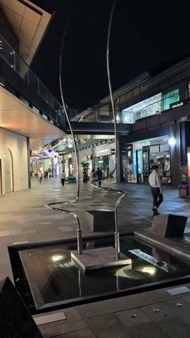 The scene is set in an outdoor shopping area at night, with store fronts visible along both sides of a pedestrian walkway. The area is paved with rectangular tiles. In the center of the frame is a water feature with a modern sculpture made of shiny metal that seems to have a water component to it. There are people walking in the distance along the walkway, some browsing store windows. A man walks along the right side of the frame. The lighting is provided by the storefronts and overhead lights, creating a well-lit environment. The sky is dark. There are modern architectural elements visible with glass railings and geometric shapes of the buildings. The sculpture is reflected in the water.