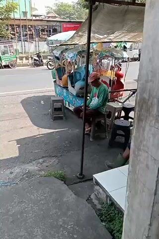 The video captures an outdoor scene next to a busy road. In the foreground, a man in a green long-sleeved shirt and a red hat sits on a stool, facing a toy ride with several colorful seats, possibly for children. The toy ride is under a canopy. To his left and right are more stools, one occupied by a person wearing green boots. In the background, vehicles, including motorcycles and cars, move along the asphalt road. Vegetation is visible along the roadside and on the right side of the frame. The overall impression is of a street-side attraction, possibly for children, with an attendant waiting for customers.