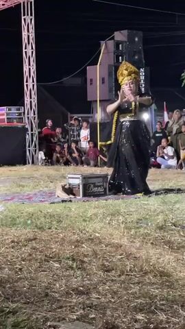 A person wearing a black sequined dress and a golden crown with black veiling is dancing on a rug placed on dry grass. The person is facing right and performing hand gestures. In the background, there is a stage with speakers and lighting equipment, and an audience of people, including children, are watching. A small wooden box with a logo that reads "DENNIS" is on the rug in front of the dancer. The scene appears to be an outdoor event at night.
