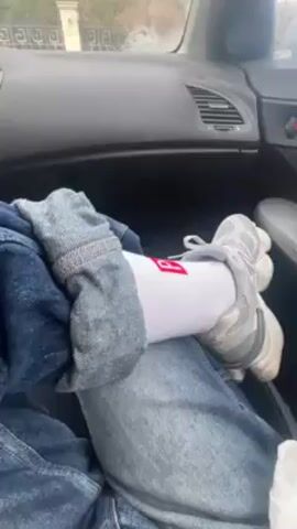 The video, taken from inside a car, features a woman in the passenger seat. She is wearing a white top, jeans, and a white jacket. She looks at the camera, pauses, and then briefly adjusts her collar. The camera angle is slightly tilted, capturing the car's interior and a blurry view of the outside through the windshield. The overall scene seems casual and uneventful.