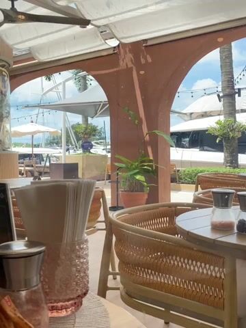The video shows a view from a restaurant table looking out towards a marina. In the foreground, there is a table with wicker chairs. On the table, there are salt and pepper shakers and a pink glass object that appears to be a vase or candle holder with napkins inside. To the left, there is a ceiling fan. Through the arches of the restaurant, we can see boats docked in the marina, umbrellas, palm trees, and a blue sky with white clouds. The overall ambiance appears to be a casual, outdoor dining experience, possibly in a warm climate.
