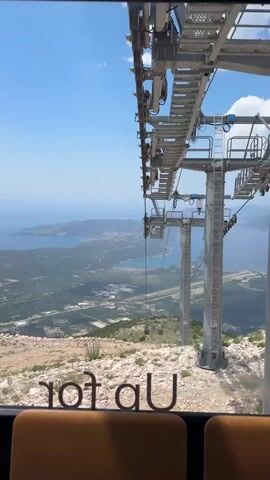 This video clip captures a scenic view from inside a cable car ascending a mountain in Kuk, Montenegro. The foreground is dominated by the interior of the cable car, with glimpses of brown upholstered seats. Through the large window, a breathtaking panoramic landscape unfolds.

The primary focus of the visual is the robust infrastructure of the cable car system. A towering metal support pylon rises impressively, with its intricate network of cables, pulleys, and maintenance platforms visible against the bright blue sky. The cable car itself is in motion, as indicated by the slightly blurred background and the presence of other cable cars suspended on the wire further down the slope.

The middle ground presents a vast expanse of the mountainous terrain of Kuk. Lush green vegetation covers the slopes, interspersed with rocky outcrops and patches of exposed earth. Below, a town or village sprawls across the valley, with buildings, roads, and some industrial or commercial structures visible. The coastline and the Adriatic Sea are prominent in the distance, with islands dotting the horizon under a clear sky with scattered white clouds.

The time of day appears to be midday, judging by the strong sunlight and the shadows cast. The weather is fair and sunny, contributing to the visibility of the expansive views. A notable detail is the text "Up for U" imprinted on the inside of the cable car window, a subtle yet effective piece of branding or a welcoming message to passengers. The overall impression is one of adventure, exploration, and the enjoyment of natural beauty from an elevated perspective.