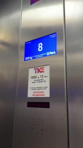 This media file documents an elevator ride, likely within a multi-story building in The Hague, Netherlands. The scene transitions between the elevator's interior panel and the exterior of the building or a different part of the elevator.

The elevator panel is the primary focus. It displays a digital screen showing the current floor as "9" and indicating a capacity of "1000 kg" and "13 Pers." Below this, a separate plaque provides information about the elevator's specifications: "1000 kg 13 Pers." It also states "Bouwjaar 2007" (Year of construction 2007) and "Install. Nr. NL0801229." A crucial warning is present: "Bij Storing de Alarmknop minimaal 3 seconden ingedrukt houden" (In case of malfunction, hold the alarm button for at least 3 seconds) and "Lift bij brand niet gebruiken" (Do not use elevator in case of fire). A red indicator light beneath this text reads "Etage Geblokkeerd" (Floor blocked).

The media then shifts to a close-up of the elevator's floor selection buttons. The button for floor "39" is illuminated with a red ring, indicating it is the current floor or has been selected. Other buttons for floors 38, 40, 35, 36, 37, 33, 34, 32, 29, 30, 31, 28, 26, 25, 22, 21, 12, 11, 13, 15, 17, 6, 5, 3, and 2 are visible. A green light is illuminated on button "38". Another section of the media shows a blurred view of what appears to be a list of floors with corresponding room numbers, likely part of the building's directory within the elevator.

The activity depicted is the functioning of an elevator, with the digital display indicating the current floor and the illuminated buttons suggesting a recent or impending floor change. The "Etage Geblokkeerd" message suggests that certain floors are inaccessible, possibly due to the blocked status. There are no people visible in the media. The setting appears to be modern and functional, typical of a commercial or residential building. The overall tone is informative, highlighting the operational status and safety instructions of the elevator.