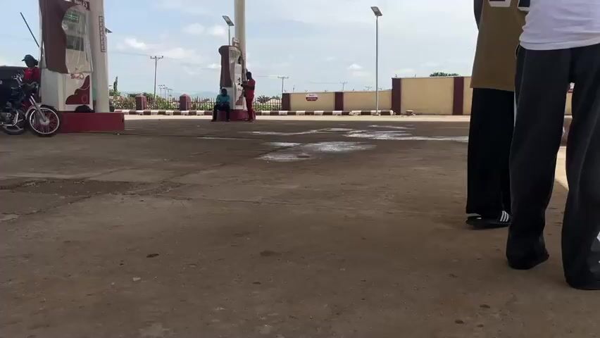 This media file captures a scene at a gas station in Pasere, Nigeria. The setting is an open-air gas station with a covered canopy supported by pillars. The ground is paved with concrete, showing some patches of wetness, suggesting recent activity or rain. The time of day appears to be daytime, with a bright, cloudy sky.

Several individuals are present, with varying activities. In the foreground, two young men are partially visible, one in a white tank top and dark pants, and another in a yellow basketball jersey with the number 24 and dark pants. They appear to be standing and observing the surroundings.

Further back, near the gas pumps, there are more people. A person in an orange shirt is seated on a motorcycle on the left. Another individual in a red outfit is standing near a gas pump. A group of people are also gathered around a motorcycle, with some seated and others standing. On the far right, another person in red is attending to a motorcycle.

The overall atmosphere seems casual, with people engaged in activities typical of a gas station. There are no strong indicators of negative emotions; the mood appears neutral to relaxed.

Visible text can be seen on the gas pumps. On the left pump, there is a logo with red and white elements and text that appears to read "Shafa." The pumps themselves have digital displays and hoses. A white barrier with red accents runs along the perimeter of the gas station. In the background, there are trees and a fence, with utility poles and lines visible, indicating a semi-urban or rural environment. A white bag with red markings lies on the ground in the middle distance.