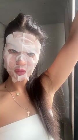 A woman with long, dark hair, pulled back from her face, is shown indoors, posing for the camera while wearing a white sheet face mask. She is wearing a white strapless top and two necklaces; one with a small, round pendant, and another with a cross pendant.

The sheet mask covers her entire face, featuring cutouts for her eyes, nose, and mouth. Throughout the video, she displays various facial expressions, including wide-eyed stares, slightly open mouths, and downturned lips, appearing to make playful or reactive faces for the camera.

She is positioned near a window, which provides a view of a city or townscape. The background features several multi-story residential buildings, some with pink and white facades, under an overcast or cloudy sky. The architecture is consistent with urban developments found in Gavrilkovo, Russia. The scene captures a casual, intimate moment of a beauty routine with an urban backdrop.