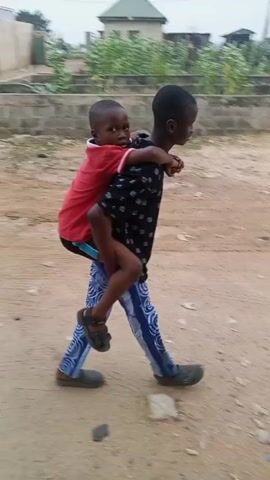 A young boy carries another younger boy on his back while walking on a dirt road. The boy doing the carrying wears a dark, patterned shirt and blue and white striped pants. The smaller boy wears a red t-shirt and is barefoot. They are walking away from the camera. The road is surrounded by trees, bushes, and buildings.