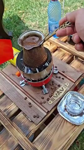 A close-up shot shows a copper pot filled with dark liquid, likely coffee, on a red camping stove. The pot is placed on a wooden pallet table outdoors, with green grass visible in the background. A hand holding a long-handled spoon stirs the liquid in the pot. Several coffee beans are scattered on the table, along with a small glass on a silver saucer and a bottle of water. A yellow and red camping gas canister is also visible to the left. The overall scene depicts the preparation of coffee in an outdoor setting.