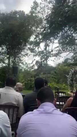 The video shows a wedding ceremony outdoors. Several people are seated in wooden chairs, facing the wedding arbor decorated with white flowers. The attendees are dressed in various formal attire, with some holding phones. The setting is a lush garden with tall trees and verdant foliage, and string lights can be seen hanging among the branches. A speaker is positioned near the arbor, presumably for amplifying the ceremony's sound.