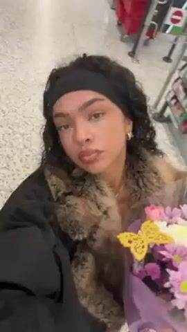 A woman is holding a bouquet of flowers and posing for a selfie. She has curly dark hair, a black headband, and hoop earrings. She wears a black jacket with a fur collar. The flowers are pink, purple, and white. She is standing on a gray tiled floor, and a merchandise rack is visible in the background. She tilts her head while holding the flowers in front of her.
